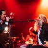 Nick Cave and Warren Ellis, two white men in black suits on stage under red lights passionately sing and gesture with outstretched arms and clenched fists.The atmosphere is bold and dramatic. Image credit: Megan Cullen