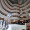 Performers in the middle of the Guggenheim, standing in a line with arms raised. Audience members stand and watch along the spiral staircase surrounding them